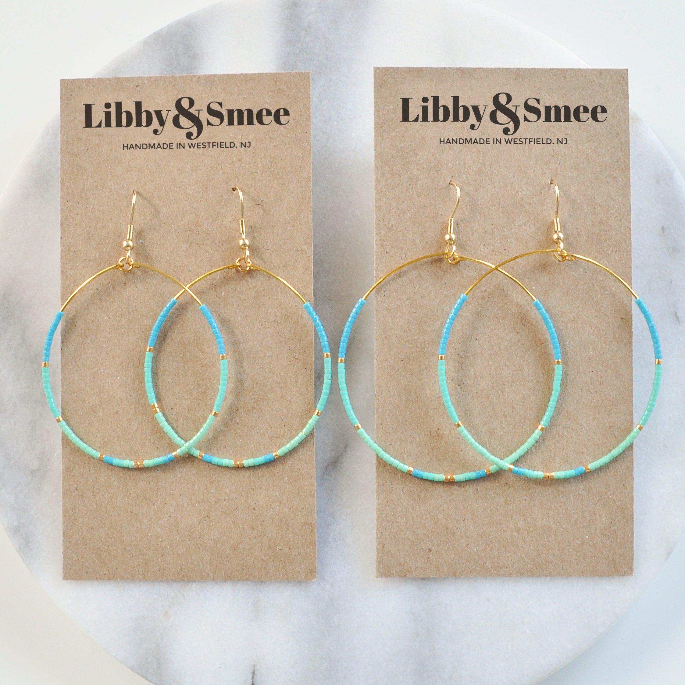 Libby & Smee Big Beaded Hoops in "Aruba" style with gold-plated ear wire and small seed beads in turquoise, aqua, mint and gold in size "big" teardrop shape and "bigger" circle shape shown on kraft earring cards with Libby & Smee Handmade in Westfield NJ logo  - still life